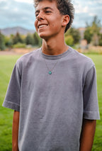 Person wearing a gray t-shirt with a necklace, standing outdoors with greenery in the background
