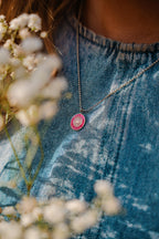 Person wearing a necklace with a pink pendant, surrounded by white flowers, on a blurred denim background.